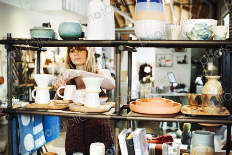 Woman standing in a shop, arranging ceramic objects on a shelf.