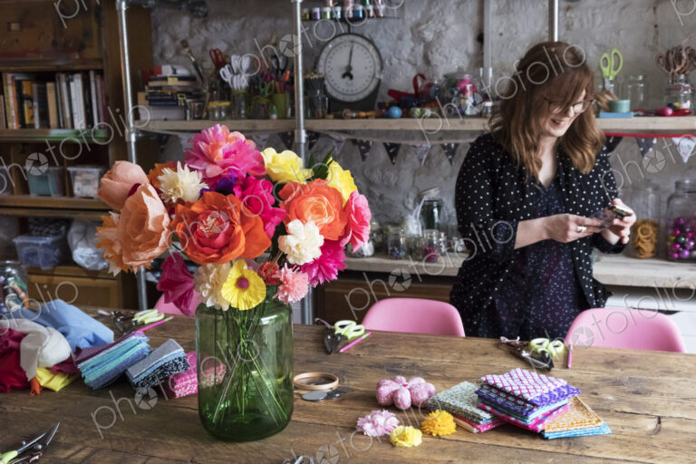 Woman standing in a workshop, bunch of colourful handcrafted fabric flowers in a vase…