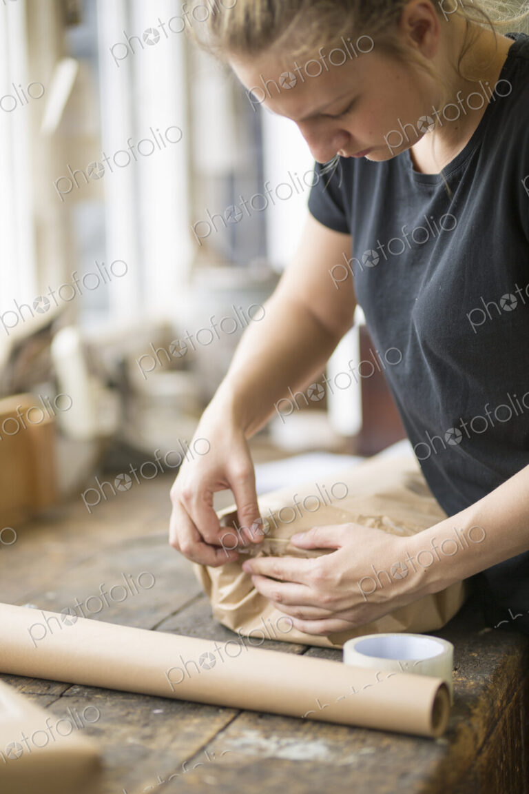Woman standing in a workshop sealing a package wrapped in brown packaging paper with…