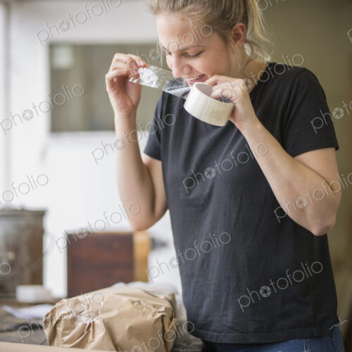 Woman standing in a workshop using her teeth on a piece of sticky tape off a roll,… Woman standing in a workshop using her teeth on a piece of sticky tape off a roll,…