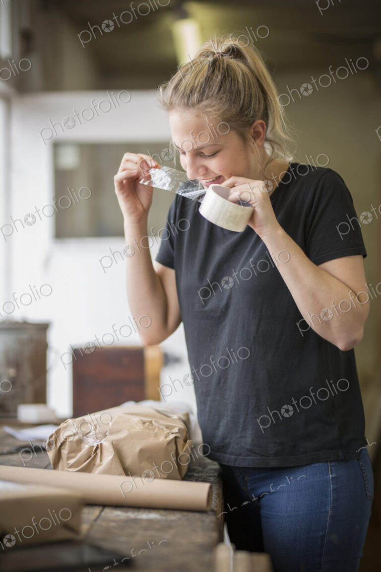 Woman standing in a workshop using her teeth on a piece of sticky tape off a roll,…