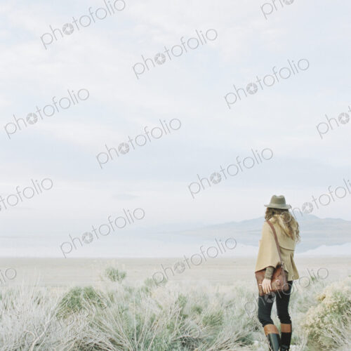 Woman standing in open ground, wearing a hat and carrying a leather bag.