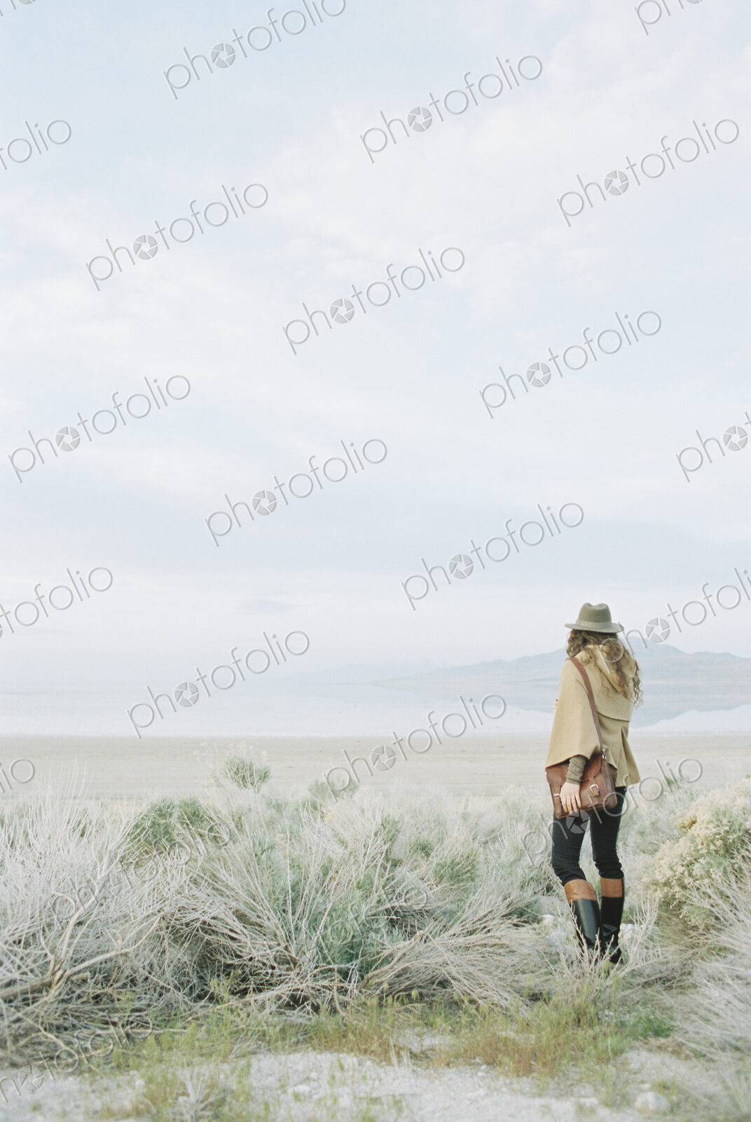 Woman standing in open ground, wearing a hat and carrying a leather bag.