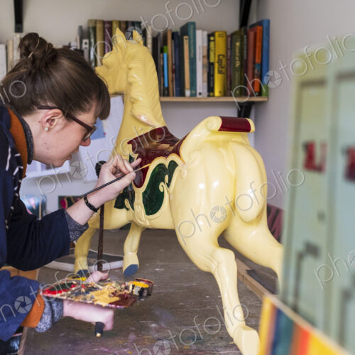 Woman standing in workshop, painting a traditional wooden carousel horse from…