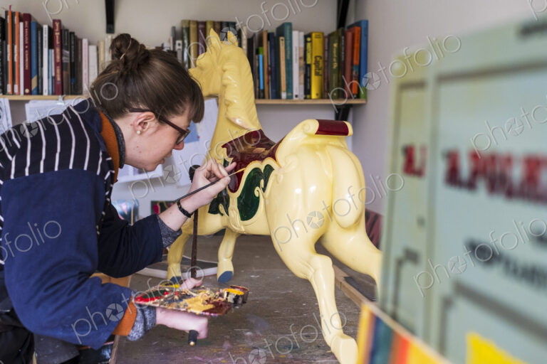 Woman standing in workshop, painting a traditional wooden carousel horse from…