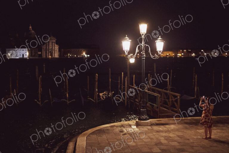 Woman taking picture of a streetlight by a lagoon in Venice, Veneto, Italy, at night.
