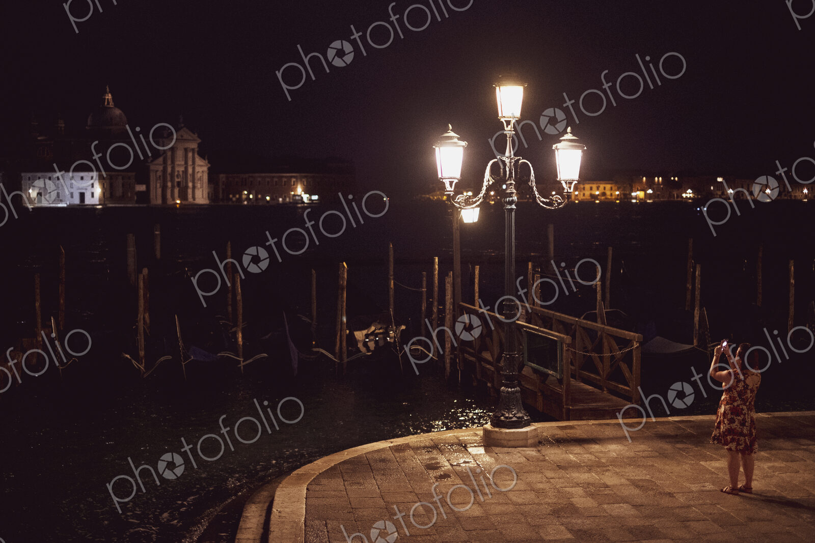 Woman taking picture of a streetlight by a lagoon in Venice, Veneto, Italy, at night.
