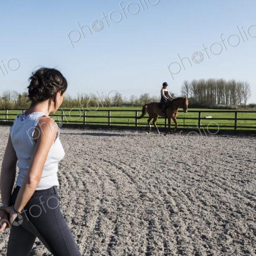 Woman trainer watching a teenage girl riding on a baybrown horse in paddock.