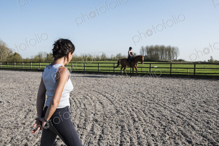 Woman trainer watching a teenage girl riding on a baybrown horse in paddock.