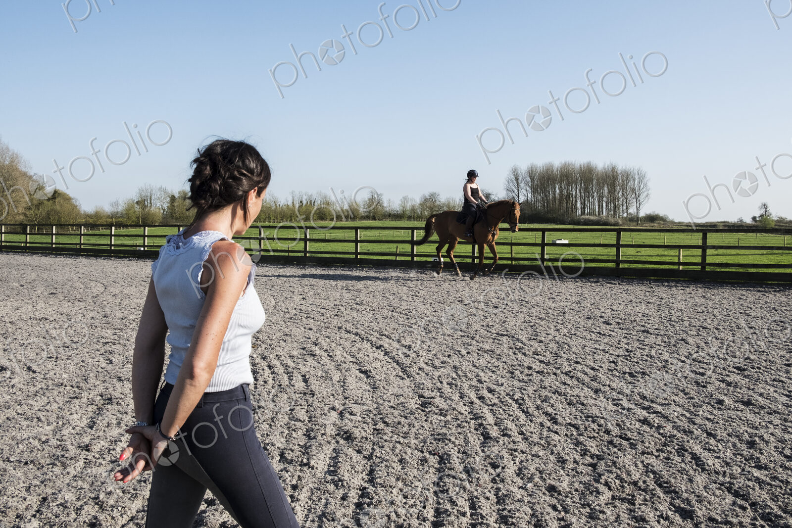 Woman trainer watching a teenage girl riding on a baybrown horse in paddock.