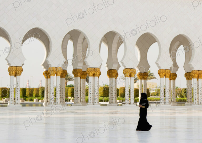Woman walking past colonnade of mosque with white washed and golden columns.