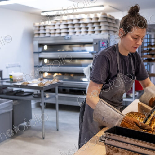 Woman wearing apron and oven gloves standing in an artisan bakery, turning out freshly…