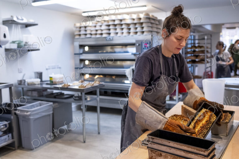 Woman wearing apron and oven gloves standing in an artisan bakery, turning out freshly…