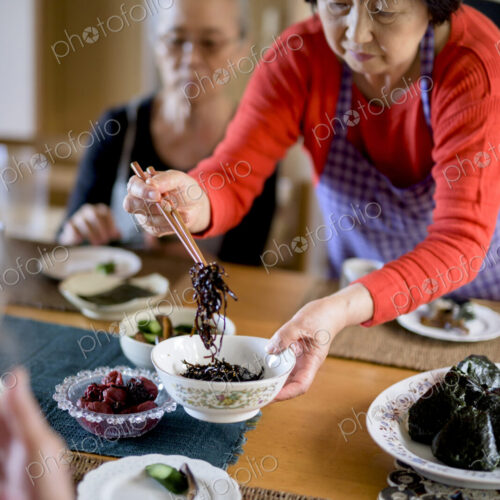 Woman wearing apron standing at kitchen table, holding bowl, serving food with chopsticks.