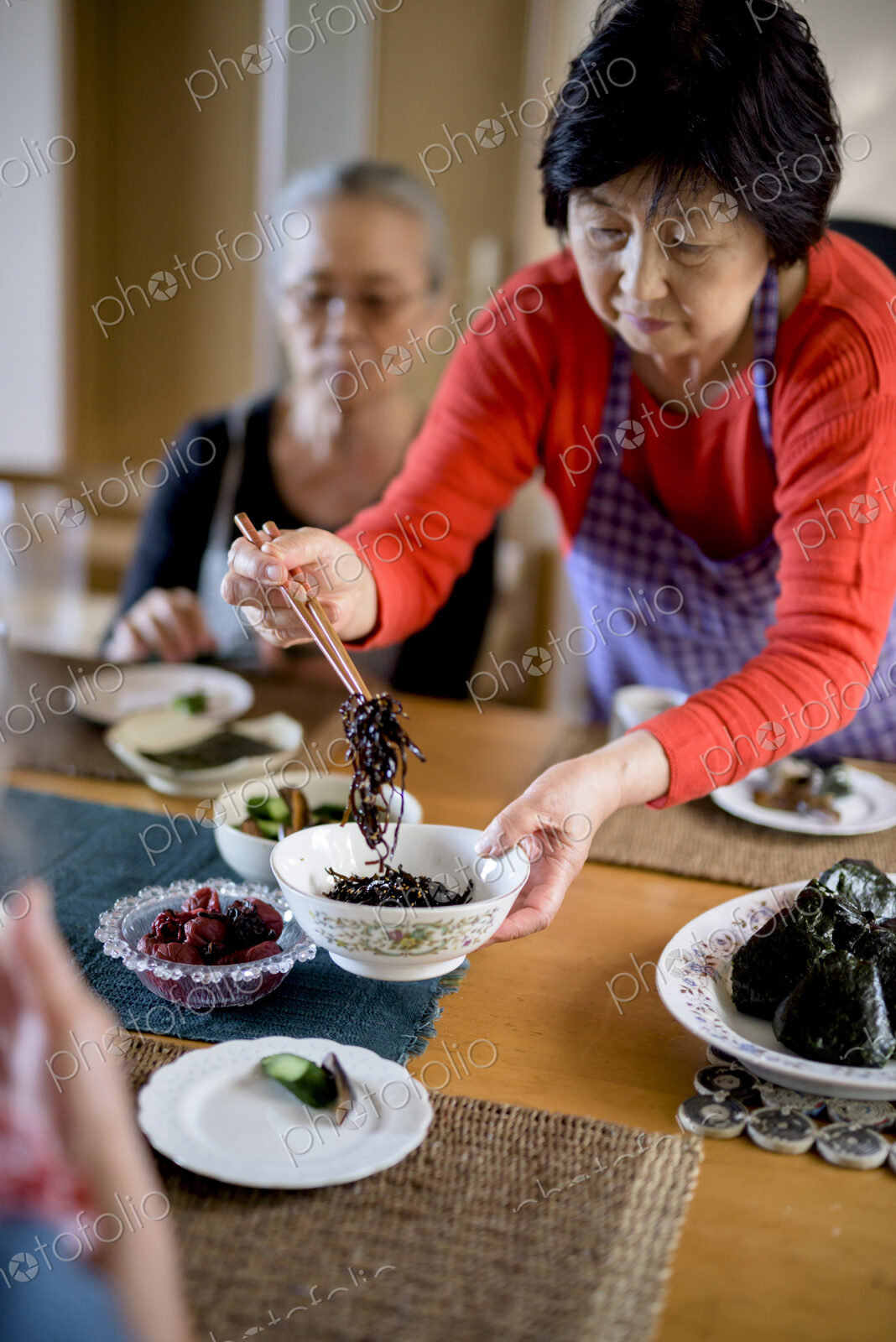Woman wearing apron standing at kitchen table, holding bowl, serving food with chopsticks.