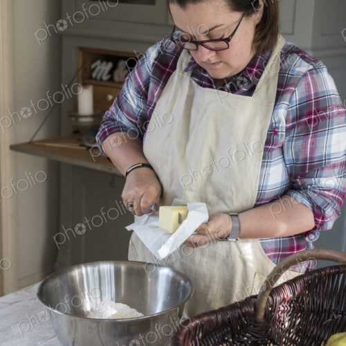 Woman wearing apron standing in a kitchen cutting butter pieces into a metal bowl… Woman wearing apron standing in a kitchen cutting butter pieces into a metal bowl…