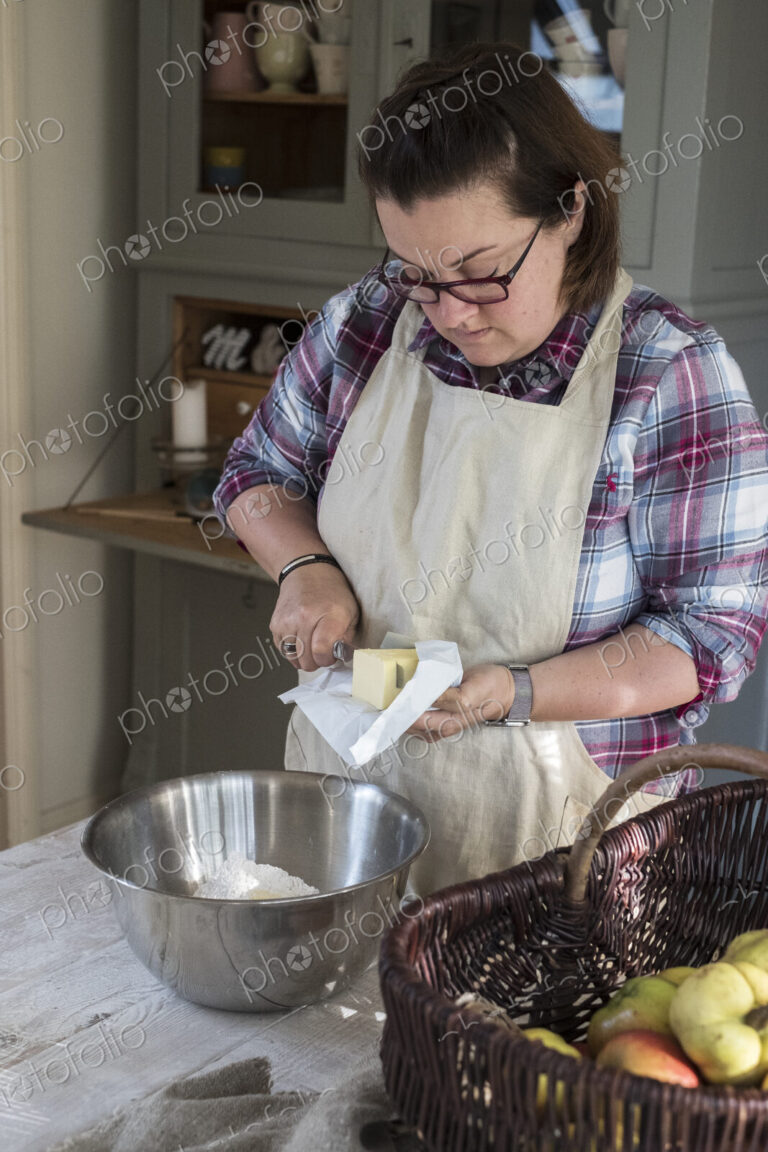 Woman wearing apron standing in a kitchen cutting butter pieces into a metal bowl…