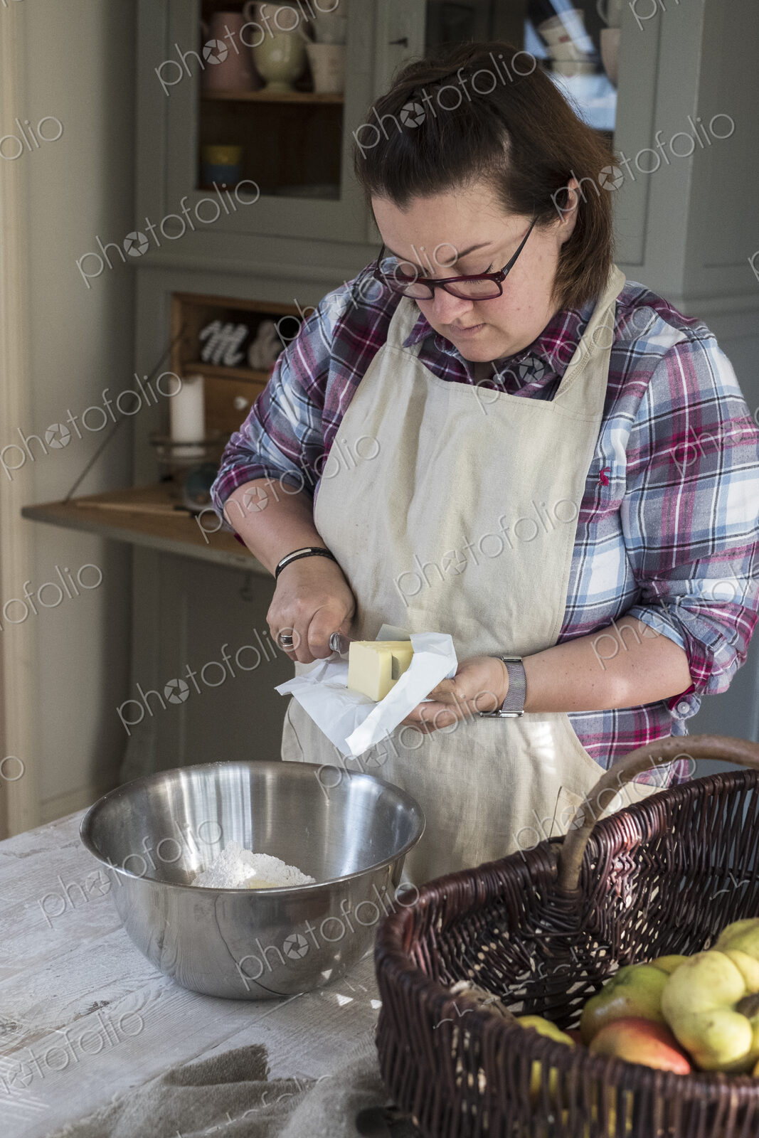 Woman wearing apron standing in a kitchen cutting butter pieces into a metal bowl…