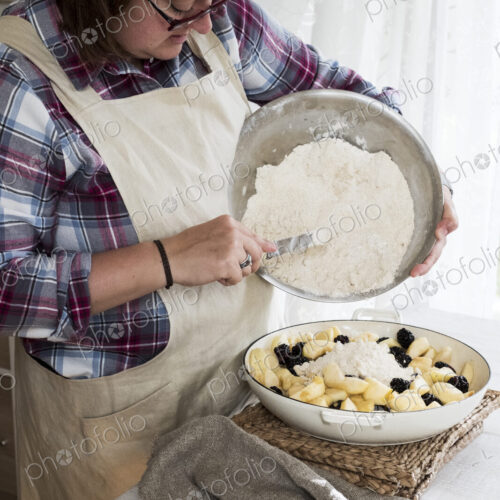 Woman wearing apron standing in a kitchen, pouring fresh crumble mixture on to a pie…