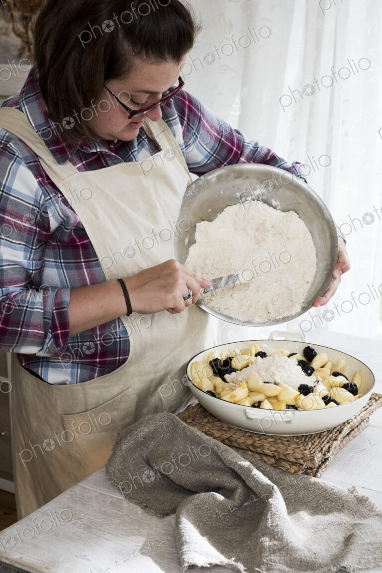 Woman wearing apron standing in a kitchen, pouring fresh crumble mixture on to a pie…