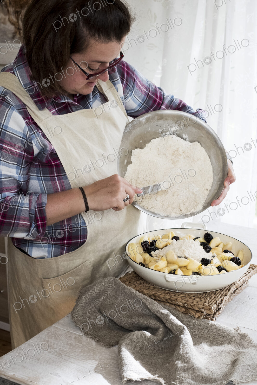 Woman wearing apron standing in a kitchen, pouring fresh crumble mixture on to a pie…