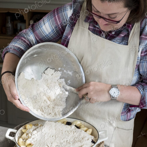 Woman wearing apron standing in a kitchen, pouring fresh crumble mixture on to a pie…
