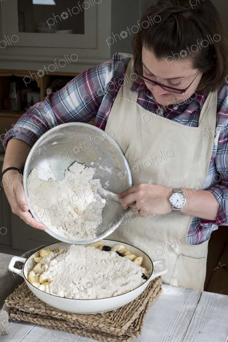 Woman wearing apron standing in a kitchen, pouring fresh crumble mixture on to a pie…
