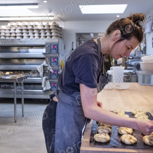 Woman wearing apron standing in an artisan bakery, brushing cinnamon buns on a baking…