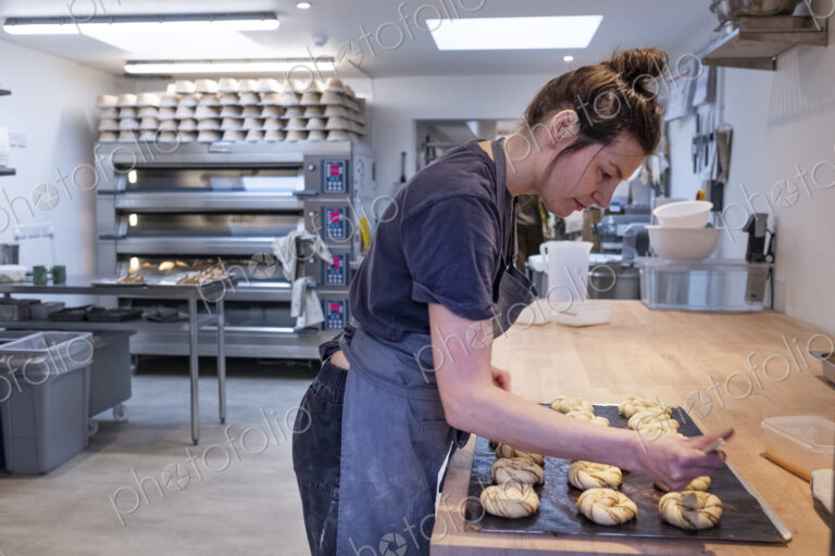 Woman wearing apron standing in an artisan bakery, brushing cinnamon buns on a baking…