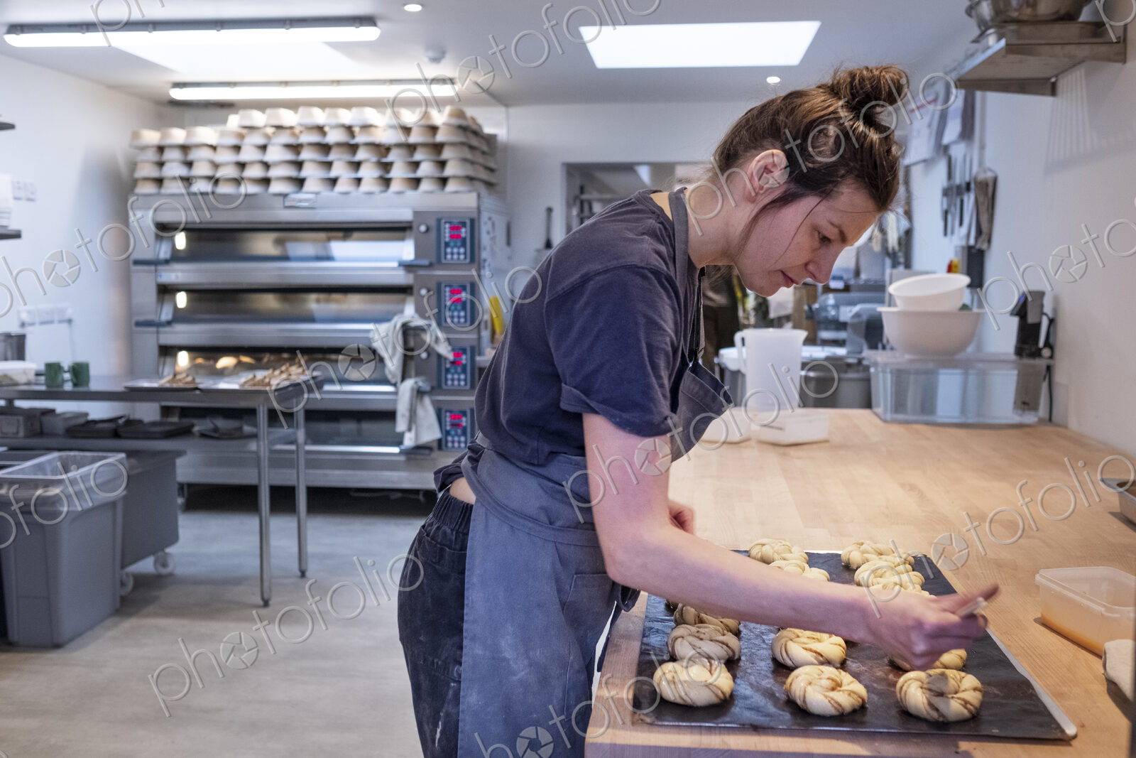 Woman wearing apron standing in an artisan bakery, brushing cinnamon buns on a baking…