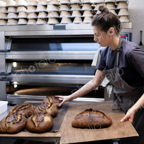 Woman wearing apron standing in an artisan bakery, placing freshly baked loaves of…