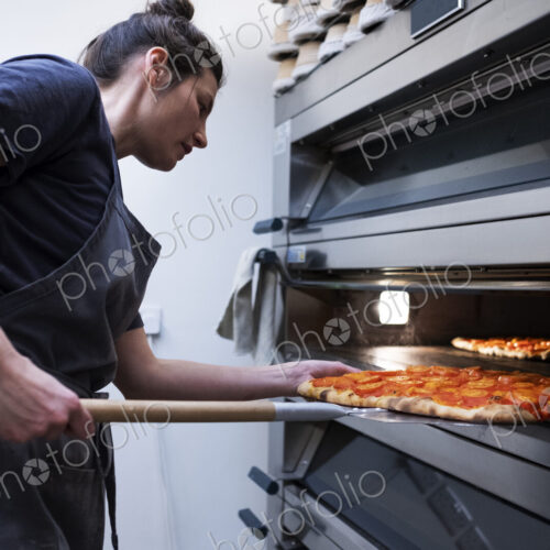 Woman wearing apron standing in an artisan bakery, placing pizza into oven.
