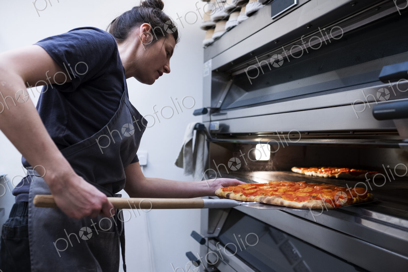 Woman wearing apron standing in an artisan bakery, placing pizza into oven.