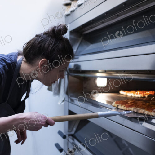 Woman wearing apron standing in an artisan bakery, placing pizza into oven.