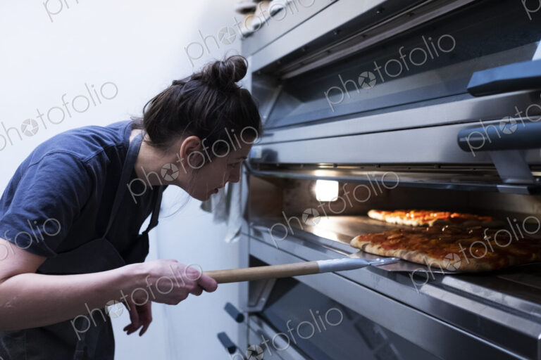 Woman wearing apron standing in an artisan bakery, placing pizza into oven.