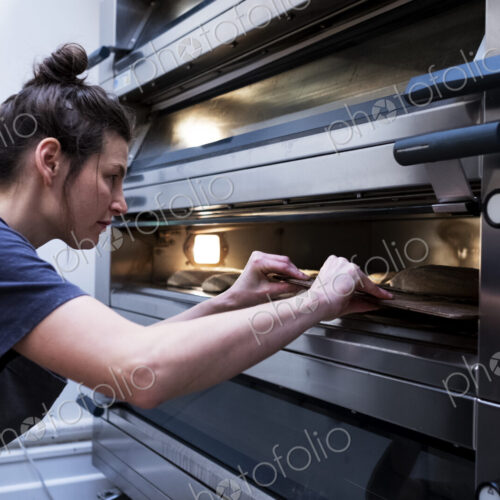 Woman wearing apron standing in an artisan bakery, placing sourdough breads into oven.