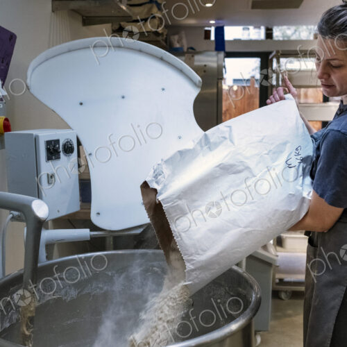 Woman wearing apron standing in an artisan bakery, pouring flour into industrial mixer.