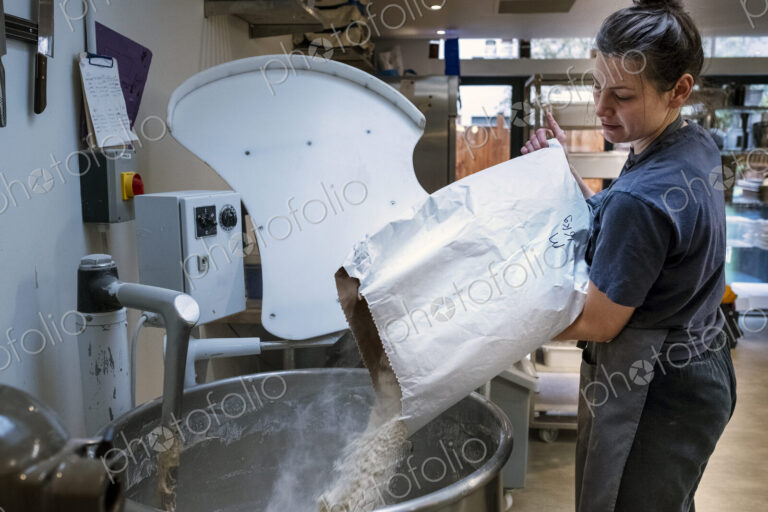 Woman wearing apron standing in an artisan bakery, pouring flour into industrial mixer.