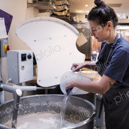 Woman wearing apron standing in an artisan bakery, pouring water into industrial mixer.