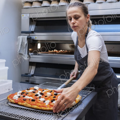 Woman wearing apron standing in an artisan bakery, preparing pizza to go into the oven.
