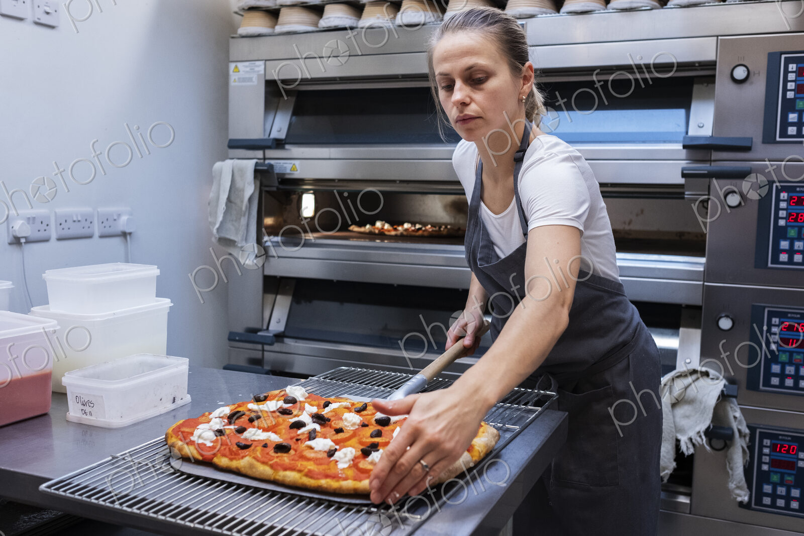 Woman wearing apron standing in an artisan bakery, preparing pizza to go into the oven.