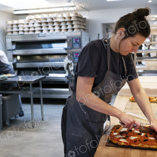 Woman wearing apron standing in an artisan bakery, slicing freshly baked pizza. Woman wearing apron standing in an artisan bakery, slicing freshly baked pizza.