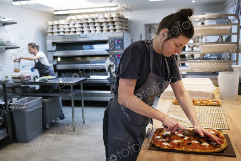 Woman wearing apron standing in an artisan bakery, slicing freshly baked pizza.