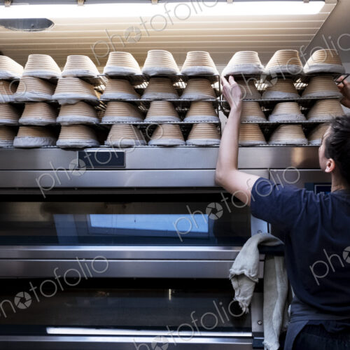 Woman wearing apron standing in an artisan bakery, stacking proving baskets on an oven. Woman wearing apron standing in an artisan bakery, stacking proving baskets on an oven.