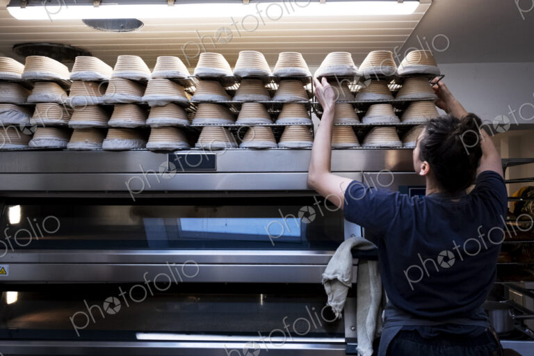 Woman wearing apron standing in an artisan bakery, stacking proving baskets on an oven.