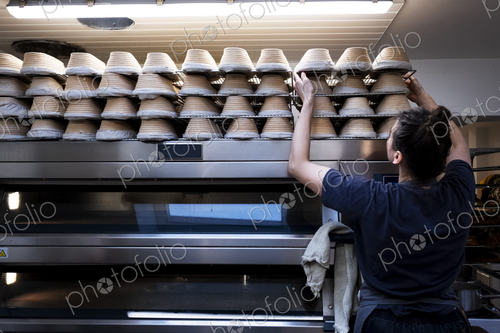 Woman wearing apron standing in an artisan bakery, stacking proving baskets on an oven.