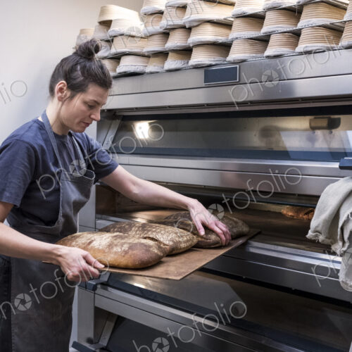 Woman wearing apron standing in an artisan bakery, taking freshly baked loaves of…