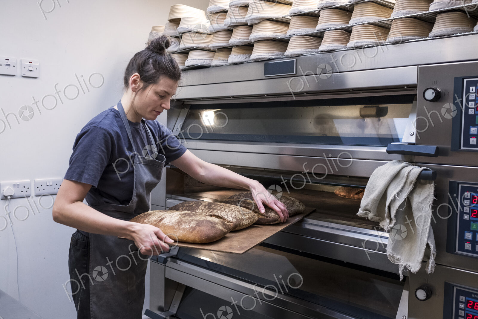 Woman wearing apron standing in an artisan bakery, taking freshly baked loaves of…