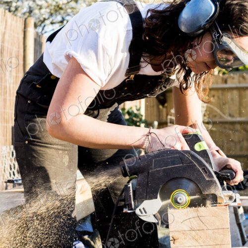 Woman wearing protective goggles and ear protectors holding circular saw, cutting… Woman wearing protective goggles and ear protectors holding circular saw, cutting…