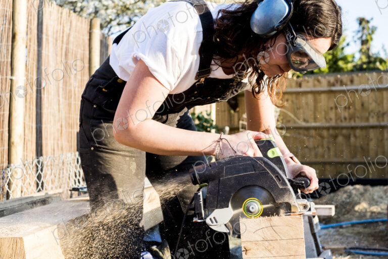 Woman wearing protective goggles and ear protectors holding circular saw, cutting…